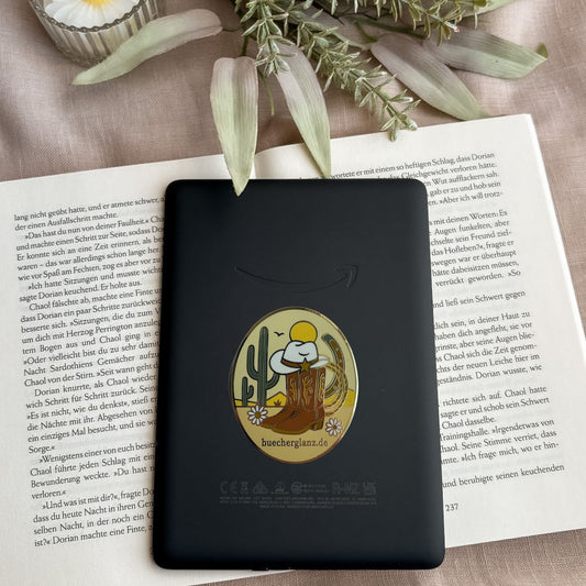 Desert Darling E-Reader Grip im Western-Design mit Cowboy-Stiefeln, Hut, Kaktus und Blumen auf schwarzem E-Reader auf offenem Buch