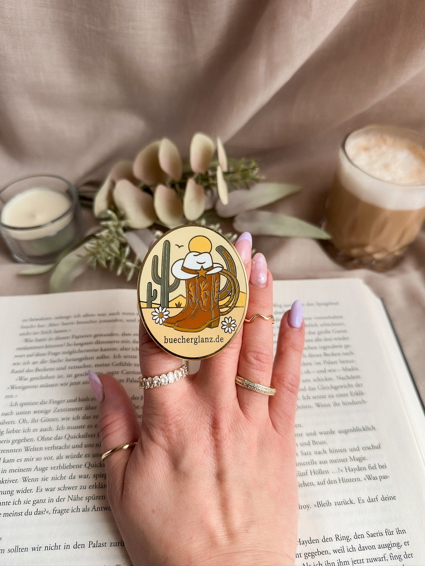 Desert Darling Kindle Grip aus Metall & Emaille – in Hand gehalten auf aufgeschlagenem Buch, Western Romance Design mit Kaktus, Cowboystiefel und Hufeisen, buecherglanz