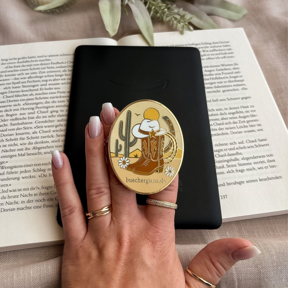Desert Darling E-Reader Grip im Western-Design mit Cowboy-Stiefeln, Hut und Blumen in der Hand vor schwarzem E-Reader