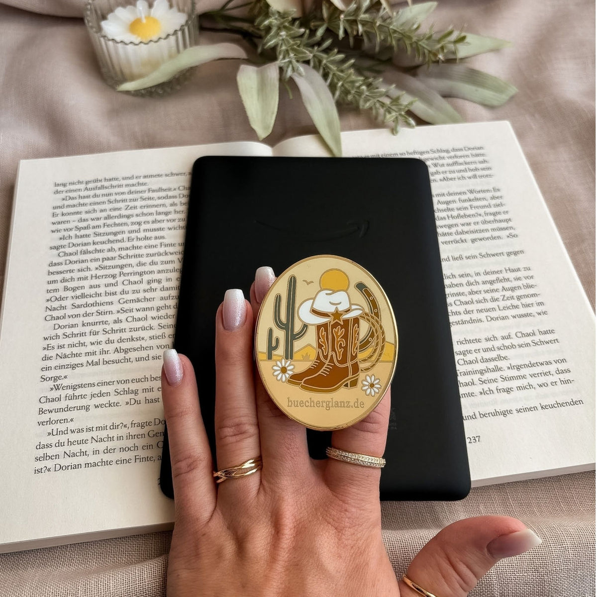 Desert Darling E-Reader Grip mit Cowboy-Stiefeln, Hut und Kaktus in der Hand vor schwarzem E-Reader auf offenem Buch