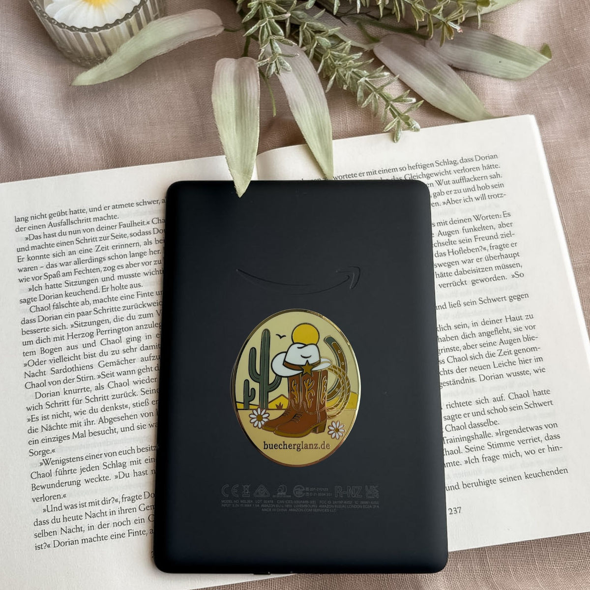 Desert Darling E-Reader Grip im Western-Design mit Cowboy-Stiefeln, Hut, Kaktus und Blumen auf schwarzem E-Reader auf offenem Buch
