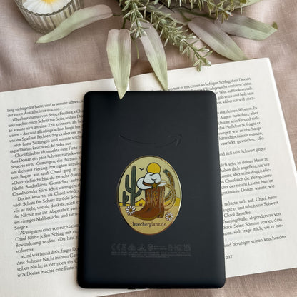 Desert Darling E-Reader Grip im Western-Design mit Cowboy-Stiefeln, Hut, Kaktus und Blumen auf schwarzem E-Reader auf offenem Buch