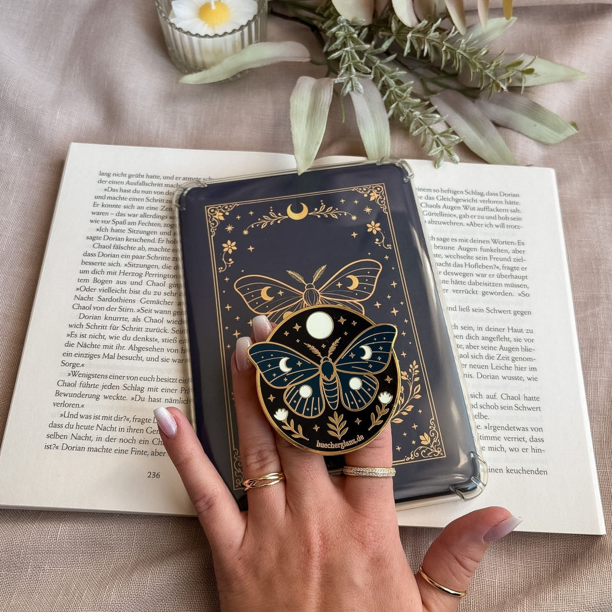 Enchanted Moth E-Reader Grip in Gold in der Hand vor passendem Insert mit Mondmotte und Mondphasen auf offenem Buch