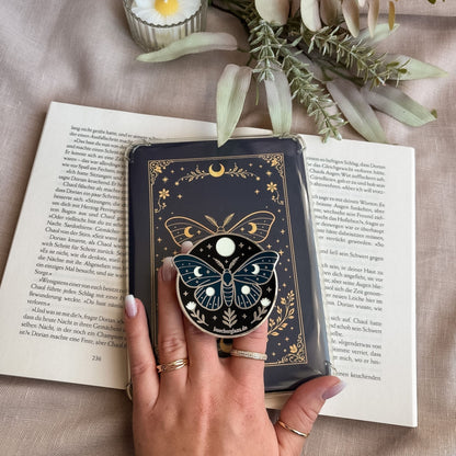 Enchanted Moth E-Reader Grip in Silber in der Hand vor passendem Insert mit Mondmotte und Mondphasen auf offenem Buch