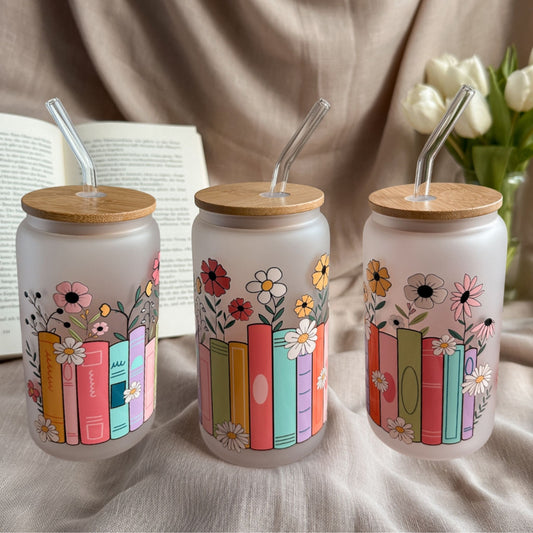 Frosted 16-oz-Glastumbler im Bookshelf-Design mit bunten Buchrücken und Wildblumen neben offenem Buch und weißen Tulpen