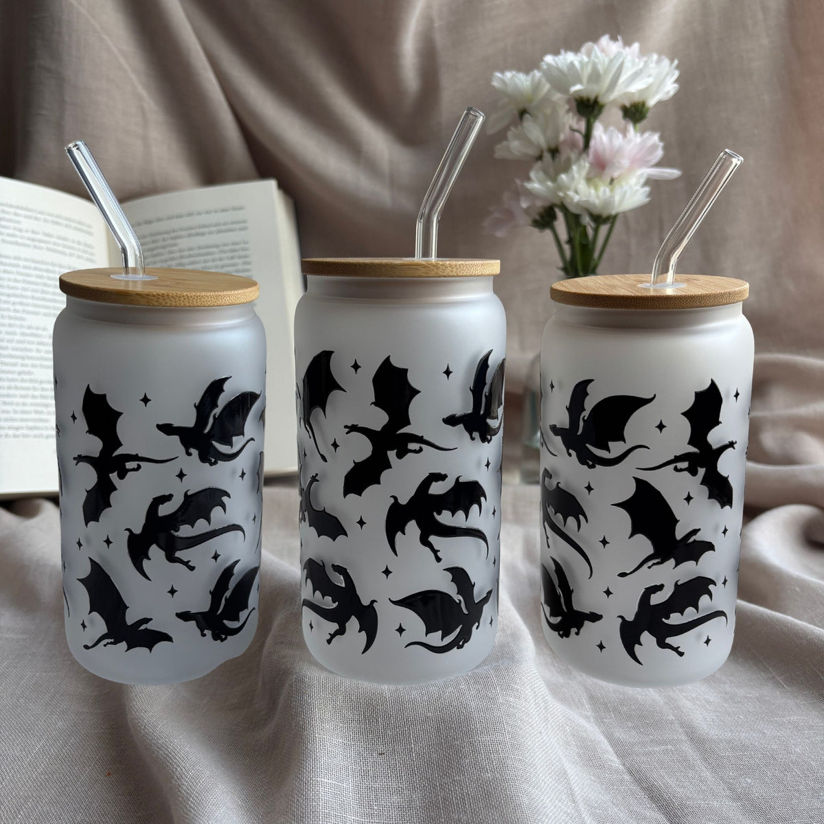 Frosted 16-oz-Glastumbler im Drachen-Design mit Bambusdeckel und Glasstrohhalm neben offenem Buch und weißen Blumen