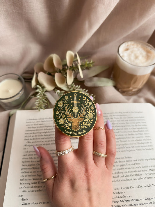 Heir of the Forest Kindle Grip Silber – in Hand gehalten auf aufgeschlagenem Buch, Romantasy Design mit Hirsch, Schwert und Blumen, Silberrand, buecherglanz