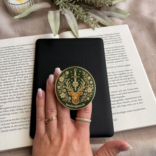 E-Reader Grip Heir of the Forest in Gold, von einer Hand an einem schwarzen E-Reader gehalten, darunter ein Buch