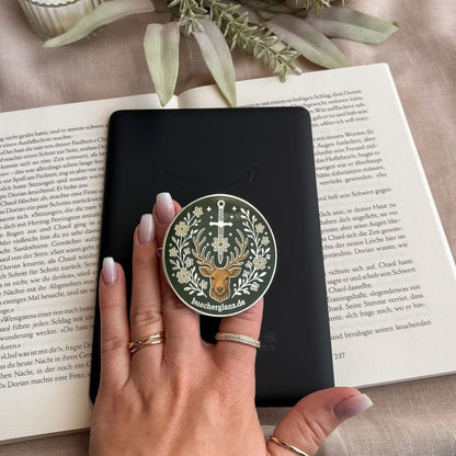 E-Reader Grip Heir of the Forest in Silber, von einer Hand an einem schwarzen E-Reader gehalten, darunter ein Buch