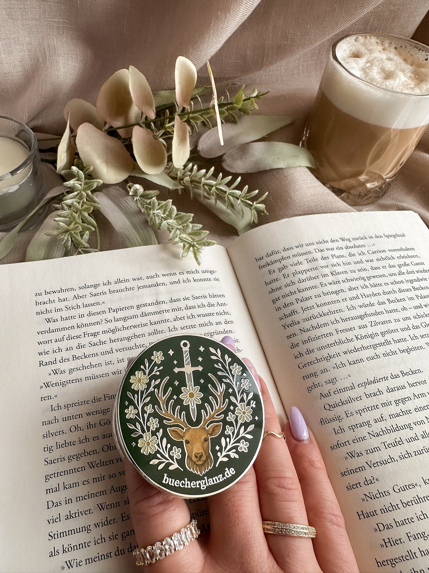 Heir of the Forest Kindle Grip Silber – in Hand gehalten auf aufgeschlagenem Buch, Romantasy Design mit Hirsch, Schwert und Blumen, Silberrand, buecherglanz