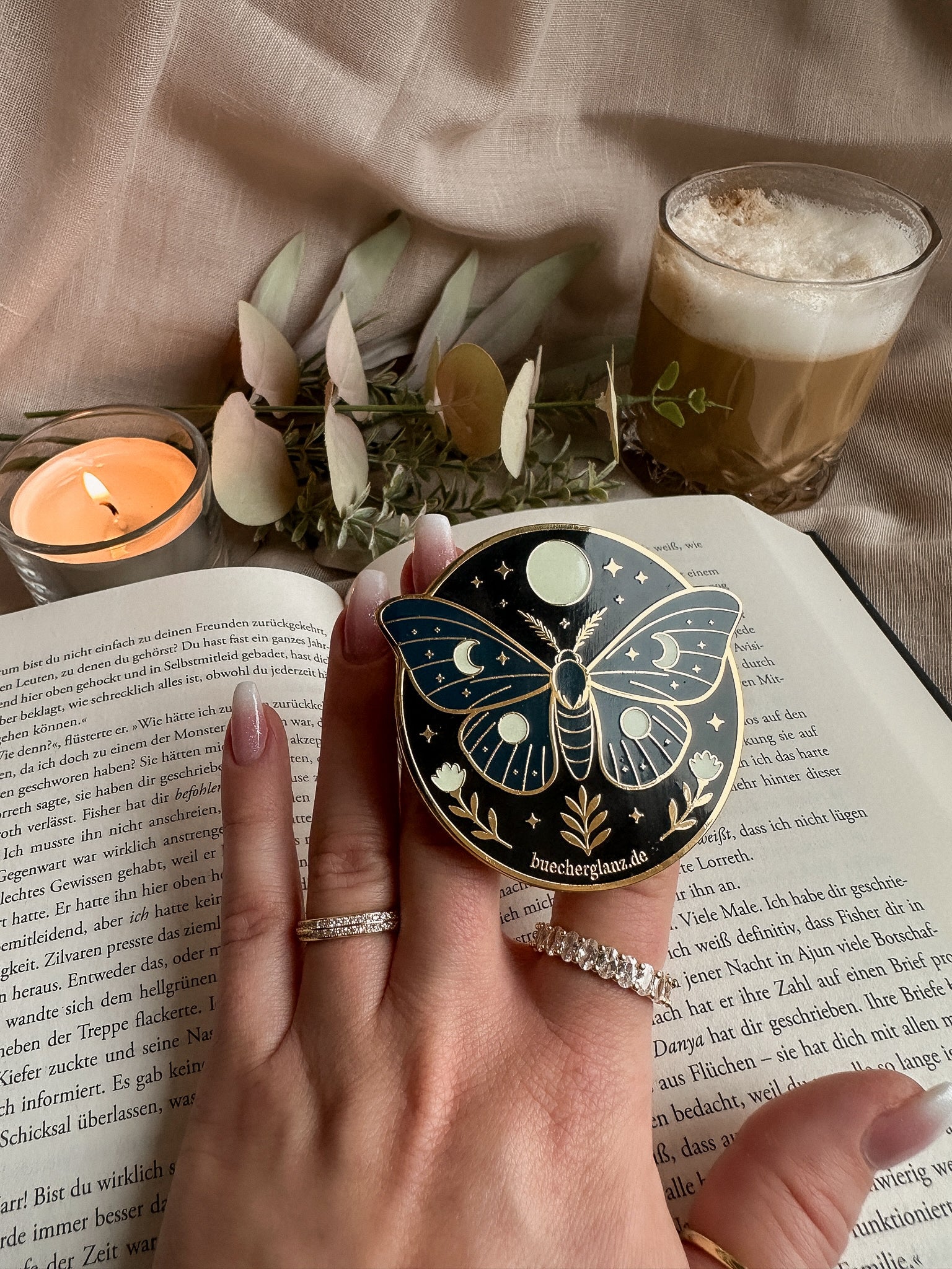 Enchanted Moth Kindle Grip in Hand auf aufgeschlagenem Buch mit Kerze und Latte
