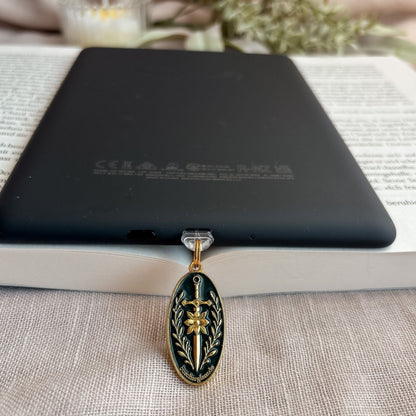 Blade & Bloom USB-C Charm Plug in Gold mit Schwert-, Blüten- und floralen Details am schwarzen E-Reader auf offenem Buch