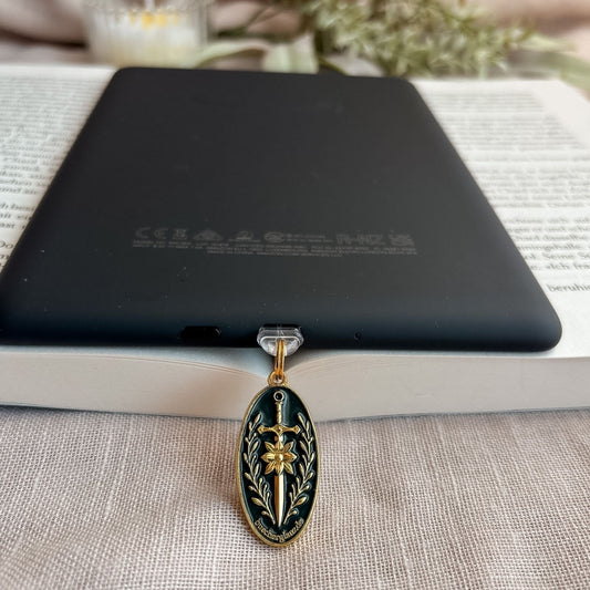 Blade & Bloom USB-C Charm Plug in Gold mit Schwert-, Blüten- und floralen Details am schwarzen E-Reader auf offenem Buch