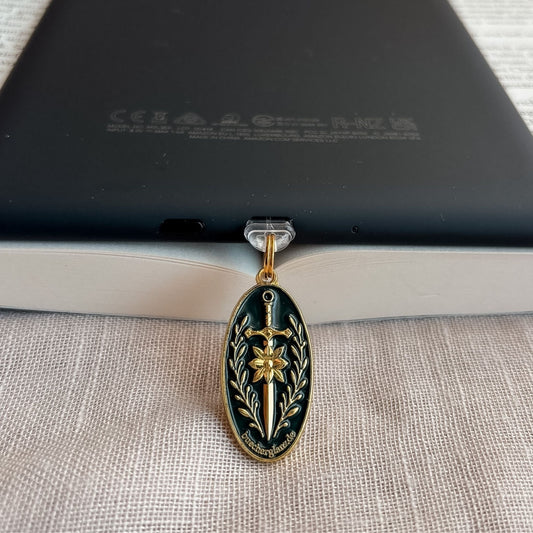 Blade & Bloom USB-C Charm Plug in Gold mit Schwert-, Blüten- und floralen Details am schwarzen E-Reader auf offenem Buch