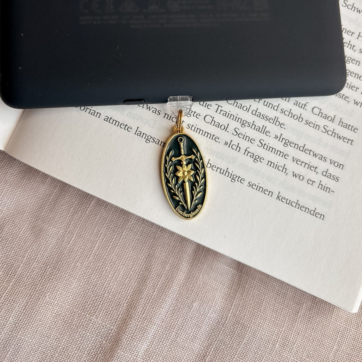 Blade & Bloom USB-C Charm Plug in Gold mit Schwertmotiv und Blütendetails am schwarzen E-Reader auf offenem Buch