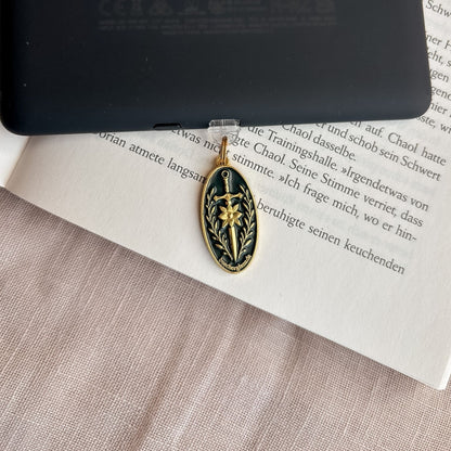 Blade & Bloom USB-C Charm Plug in Gold mit Schwertmotiv und Blütendetails am schwarzen E-Reader auf offenem Buch